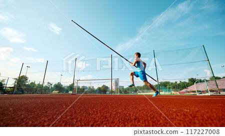 High jump training in progress, low angle view on competitive young man, pole jump athlete with pole in motion at outdoor track. 117227008
