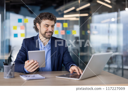 Confident businessman working with digital tablet and laptop in modern office. Professional wearing formal attire, focusing on work tasks, surrounded by sticky notes on glass walls 117227009