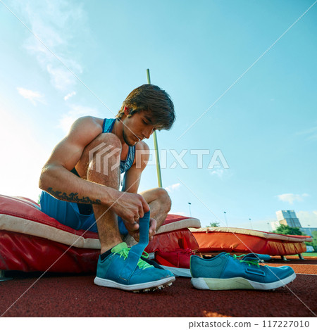 Muscular young men, pole vault athlete sitting at outdoor stadium and getting ready for enduring training session under clear blue sky 117227010