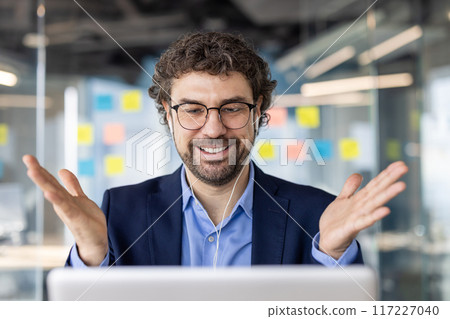 Confident businessman in office engaged in virtual meeting, smiling and gesturing while discussing ideas and strategies, 117227040