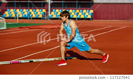 Young man in blue uniform, pole vault athlete doing warming up exercises on sport track, outdoor stadium before training session 117227041