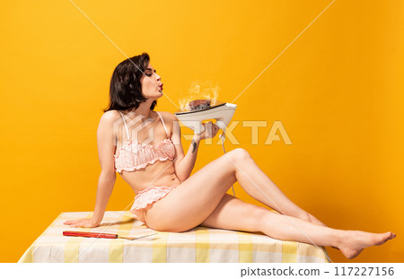 Woman in pink lace lingerie sits on table, blowing on sizzling burger cooking on iron against bright yellow background. Woman in pink lace lingerie sits on table, blowing on sizzling burger cooking on iron against bright yellow background. 117227156