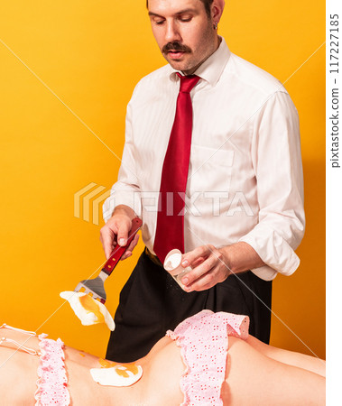 Playful photo of man in white shirt and red tie salts fried egg on woman's back, who wearing pink lace lingerie against yellow background. 117227185
