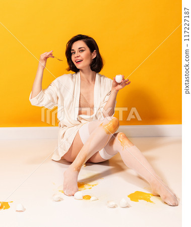 Woman, smiling playfully, in white robe and stockings sits on floor, holding egg and spoon, with broken eggs and yolk around her against yellow background. Woman, smiling playfully, in white robe and stockings sits on floor, holding egg and spoon, with broken eggs and yolk around her against yellow background. 117227187
