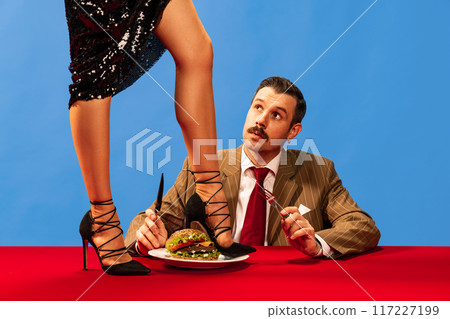 Stylish woman in heels pours liquid into glass from above, while man in suit sits at red table with burger, looking up in surprise. 117227199