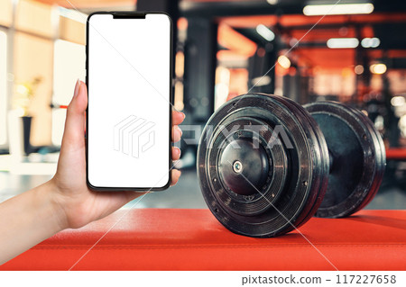 Dumbbells close-up in the gym. Hand holding a smartphone with mock up. Copy space. Concept of fitness Dumbbells close-up in the gym. Hand holding a smartphone with mock up. Copy space. Concept of fitness 117227658