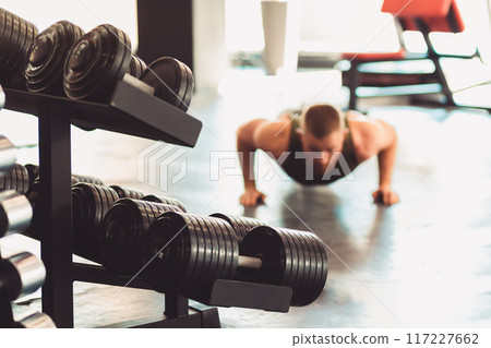 Blurred man does push-ups on the gym floor on the background. Dumbbells on the foreground. Concept of sports and warm-up 117227662
