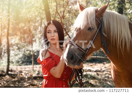 Portrait of Young beautiful brunette woman in red dress posing near horse. Sunlight Portrait of Young beautiful brunette woman in red dress posing near horse. Sunlight 117227711