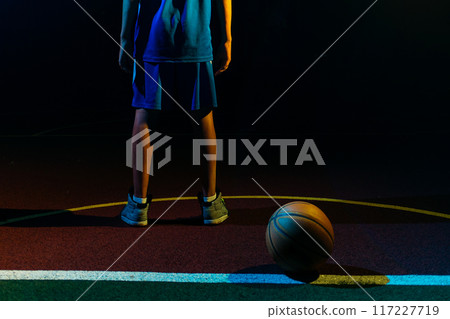 Basketball. A teenage boy in a blue sportswear stands on the Playground with ball. Close up of legs. Black background. Rear view. Concept of sports games Basketball. A teenage boy in a blue sportswear stands on the Playground with ball. Close up of legs. Black background. Rear view. Concept of sports games 117227719
