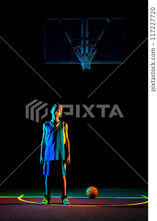 Basketball. A teenager in blue sportswear poses on the Playground, next to a basketball. Dark background, multi-colored neon lighting. Vertical. Copy space. Concept of sports games Basketball. A teenager in blue sportswear poses on the Playground, next to a basketball. Dark background, multi-colored neon lighting. Vertical. Copy space. Concept of sports games 117227720