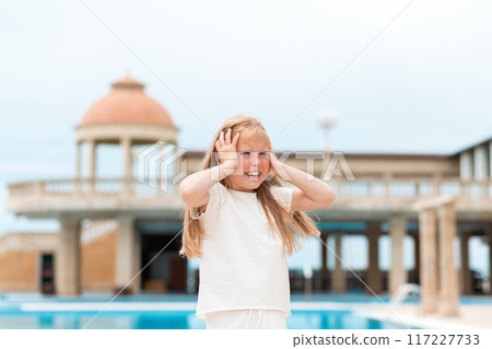 Emotions. Portrait of a frightened crying little girl holding her head. Headache. In the background there is a building with a dome and a swimming pool 117227733