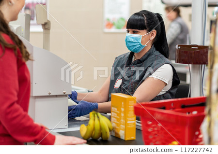 A young woman in a medical mask and gloves, working positively at the checkout in a supermarket. Concept of coronovirus, protection from infection and industrial crisis A young woman in a medical mask and gloves, working positively at the checkout in a supermarket. Concept of coronovirus, protection from infection and industrial crisis 117227742