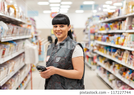 A young smiling Caucasian woman who works as a Manager in a supermarket, posing between the food rows, in the aisle. Business and technology concepts A young smiling Caucasian woman who works as a Manager in a supermarket, posing between the food rows, in the aisle. Business and technology concepts 117227744