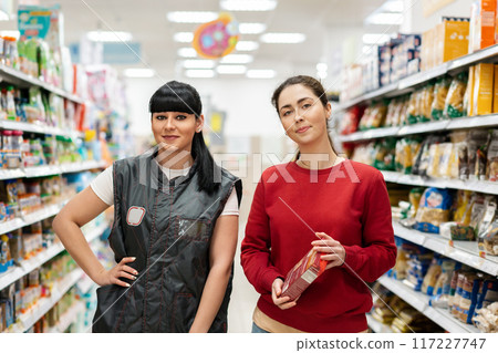 Store employees, the Director and Manager, pose against the background of food shelves, in the aisle. Two beautyful young Caucasian women. Concept of shopping, business and trade Store employees, the Director and Manager, pose against the background of food shelves, in the aisle. Two beautyful young Caucasian women. Concept of shopping, business and trade 117227747