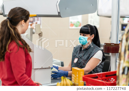 A young woman in a medical mask and gloves, working at the checkout in a supermarket. In the foreground is a customer in a blur. Concept of coronovirus, protection from infection and industrial crisis A young woman in a medical mask and gloves, working at the checkout in a supermarket. In the foreground is a customer in a blur. Concept of coronovirus, protection from infection and industrial crisis 117227748