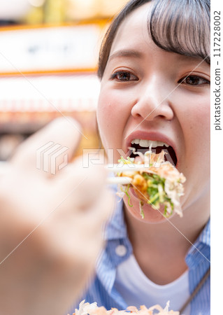 A woman eating takoyaki outdoors in bright light in Dotonbori, Osaka, Osaka Prefecture 117228002