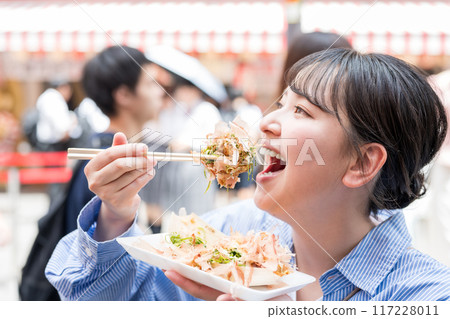 A woman eating takoyaki outdoors in bright light in Dotonbori, Osaka, Osaka Prefecture 117228011
