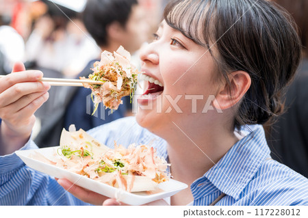 A woman eating takoyaki outdoors in bright light in Dotonbori, Osaka, Osaka Prefecture 117228012