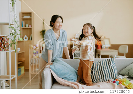 Asian little girl playing on sofa and looking directly at camera while her mother smiling and watching her in light nursery Asian little girl playing on sofa and looking directly at camera while her mother smiling and watching her in light nursery 117228045