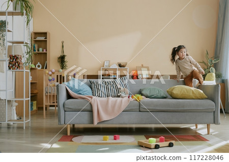 Cute little Asian girl fooling around on soft gray sofa in light beige nursery equipped with toys and furniture 117228046