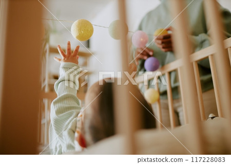 Low angle shot of infant lying in baby cot and playing with multicolored rattles while his mother standing next to cot Low angle shot of infant lying in baby cot and playing with multicolored rattles while his mother standing next to cot 117228083