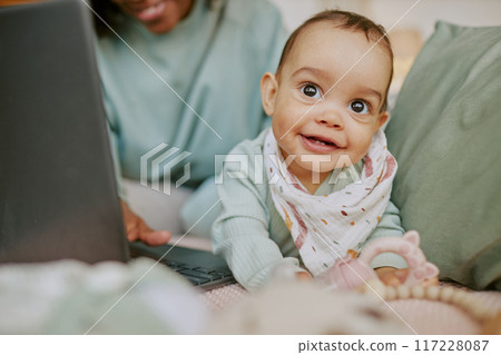 Baby boy wearing flax bib fooling around on comfortable bed while his mother working remotely via laptop in background 117228087