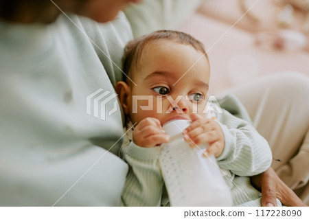 Medium close up of male infant holding baby bottle with formula and eating by himself while his mother holding him 117228090