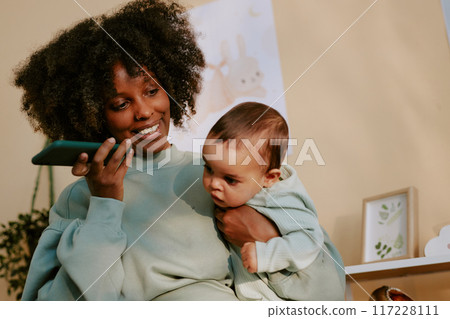 Smiling African American mom holding her baby boy while listening to voice message via phone 117228111