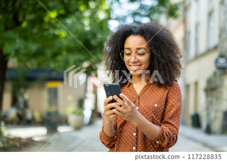 Smiling woman using smartphone while standing on a city street. Bright sunny day with green trees in the background. Urban lifestyle and communication concept. Smiling woman using smartphone while standing on a city street. Bright sunny day with green trees in the background. Urban lifestyle and communication concept. 117228835