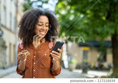 A cheerful woman tourist is seen strolling through a city, enjoying her vacation. She is holding a credit card and using her mobile phone, embodying both relaxation and modern lifestyle. A cheerful woman tourist is seen strolling through a city, enjoying her vacation. She is holding a credit card and using her mobile phone, embodying both relaxation and modern lifestyle. 117228839