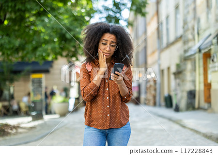 Woman with worried expression looking at phone while walking outdoors in an urban environment. Concept of concern, unexpected news, or social media issue. 117228843