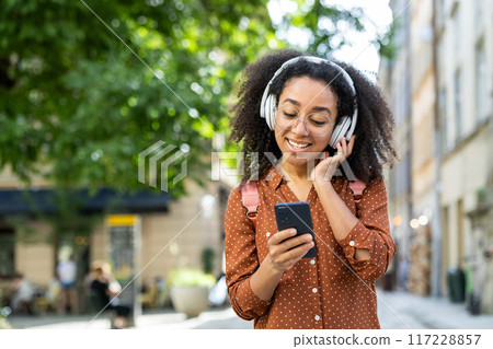 Smiling woman enjoying music on headphones while using smartphone in city street. Relaxed and happy, she is immersed in her device in outdoor setting. Smiling woman enjoying music on headphones while using smartphone in city street. Relaxed and happy, she is immersed in her device in outdoor setting. 117228857