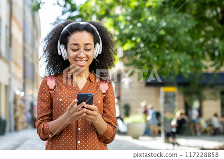 Woman tourist wearing headphones and using a smartphone while strolling through the city on vacation. The image captures a sense of joy and relaxation as she enjoys her surroundings. Woman tourist wearing headphones and using a smartphone while strolling through the city on vacation. The image captures a sense of joy and relaxation as she enjoys her surroundings. 117228858
