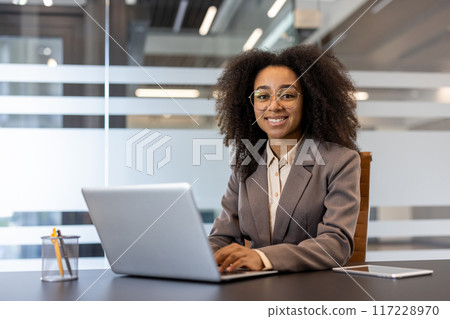 Smiling businesswoman wearing glasses and a suit, working on a laptop at a desk in a modern office. The background features glass panels and a professional atmosphere. 117228970