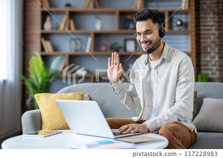 Young professional man working from home on laptop during video conference call. Smiling and waving while sitting in comfortable living room, wearing headset with microphone 117228983