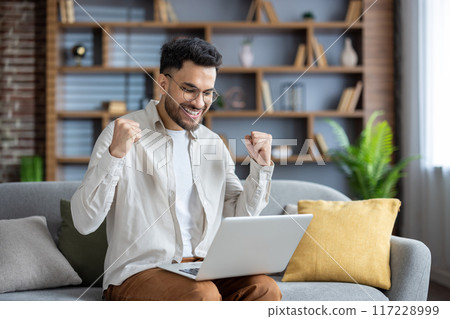 Young man sitting on couch using laptop, expressing joy and excitement. Celebrates positive achievement, success, or good news. Modern home setting with bookshelf and plants in background. 117228999