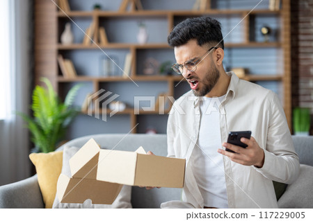 Frustrated man opening delivery box at home while holding smartphone, feeling disappointed with order. Concept of online shopping issues, customer service, dissatisfaction, and negative emotions Frustrated man opening delivery box at home while holding smartphone, feeling disappointed with order. Concept of online shopping issues, customer service, dissatisfaction, and negative emotions 117229005