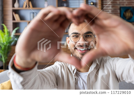 Happy young man with glasses making heart shape with his hands while looking at camera during video call at home, showing love and connection. Cozy living room background with bookshelf and plants. 117229010