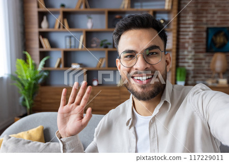 Young man waving and smiling during video call using phone at home. Casual and friendly setting with bookshelf in background. Concept of remote work, communication, and connection. Young man waving and smiling during video call using phone at home. Casual and friendly setting with bookshelf in background. Concept of remote work, communication, and connection. 117229011