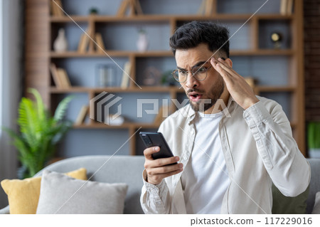 Young man in glasses looking surprised and shocked at his smartphone while sitting on couch at home. Expressing emotions, disbelief, and astonishment. Modern casual lifestyle concept. Young man in glasses looking surprised and shocked at his smartphone while sitting on couch at home. Expressing emotions, disbelief, and astonishment. Modern casual lifestyle concept. 117229016