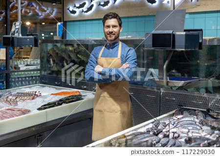 Confident fishmonger standing with arms crossed in front of seafood display at fish market. Fresh fish and seafood in foreground. Man wearing apron smiling at camera. Confident fishmonger standing with arms crossed in front of seafood display at fish market. Fresh fish and seafood in foreground. Man wearing apron smiling at camera. 117229030