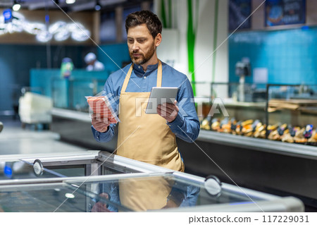 Supermarket employee wearing apron using tablet checking stock in grocery store. Man holding tablet and frozen food in supermarket. Retail worker checking inventory ensuring product availability Supermarket employee wearing apron using tablet checking stock in grocery store. Man holding tablet and frozen food in supermarket. Retail worker checking inventory ensuring product availability 117229031