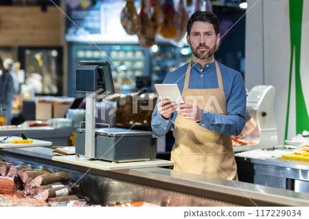 Male butcher in apron using tablet behind counter at deli shop. Display of various meats and sausages visible. Digital transformation in traditional retail setting 117229034
