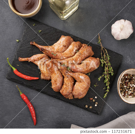Delicacies. Grilled BBQ quail legs in marinade and spices on black slate with fresh herbs and sauce Delicacies. Grilled BBQ quail legs in marinade and spices on black slate with fresh herbs and sauce 117231671