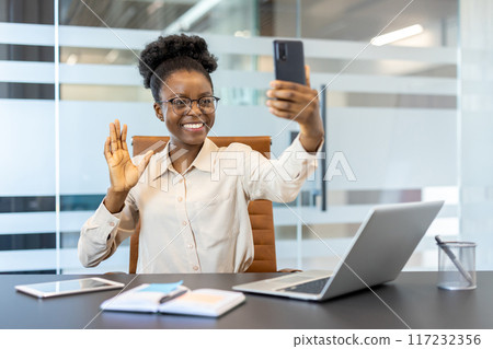 Young african american businesswoman using phone for video call, smiling and waving at workplace inside modern office with laptop and documents Young african american businesswoman using phone for video call, smiling and waving at workplace inside modern office with laptop and documents 117232356