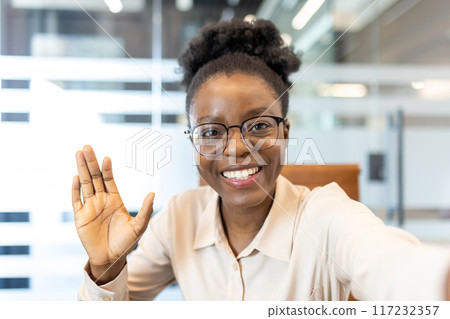 Smiling woman with glasses waving at camera during video call in office. Happy professional greeting while looking at the camera using phone. Concept of virtual meetings and remote communication. Smiling woman with glasses waving at camera during video call in office. Happy professional greeting while looking at the camera using phone. Concept of virtual meetings and remote communication. 117232357
