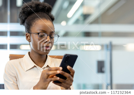 Young african american businesswoman using phone at workplace inside office. Professional woman focused on screen, wearing glasses, corporate attire. Modern business represents productivity Young african american businesswoman using phone at workplace inside office. Professional woman focused on screen, wearing glasses, corporate attire. Modern business represents productivity 117232364