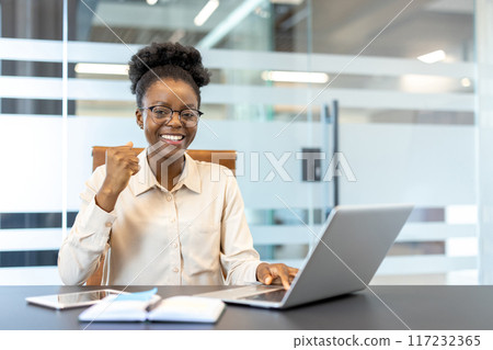 Young African American businesswoman in office using laptop, smiling and showing success. Professional work environment with modern technology and determined expression Young African American businesswoman in office using laptop, smiling and showing success. Professional work environment with modern technology and determined expression 117232365