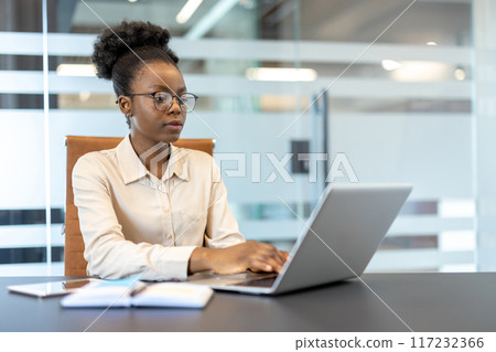 Young african american businesswoman focused while working on laptop at workplace in modern office setting. Serious and concentrated expression showcasing productivity and professionalism. Young african american businesswoman focused while working on laptop at workplace in modern office setting. Serious and concentrated expression showcasing productivity and professionalism. 117232366