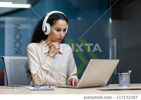 Focused woman wearing headphones working on laptop in modern office. Professional concentrating on task, using computer and technology. Concept of remote work, business, productivity 117232387
